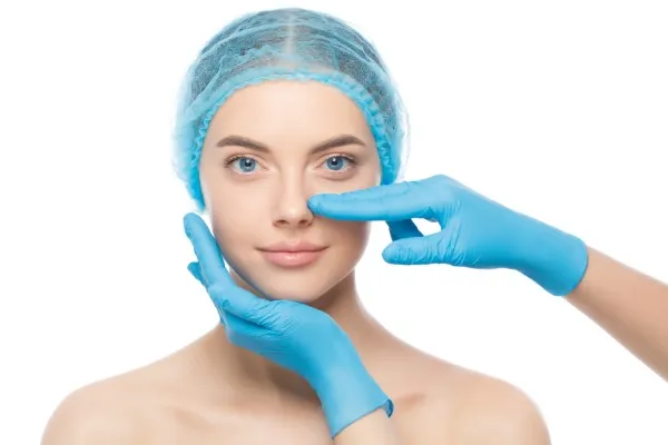 Female patient wearing a surgical cap during a plastic surgery consultation, with doctor’s hands gently analyzing her nose before rhinoplasty