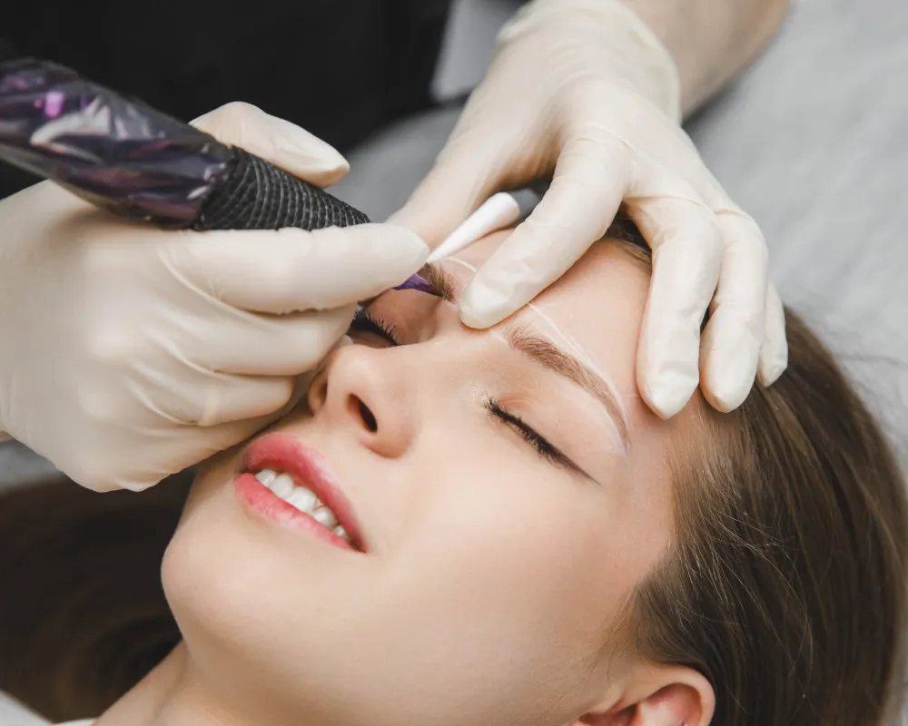 A professional performing a microblading procedure on a female client’s eyebrows using a manual tool to create hair-like strokes in the context of microblading vs micropigmentation