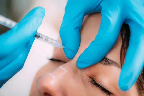 A focused shot of a medical professional carefully administering a cosmetic injectable near the patient's eyebrow, marked for precision