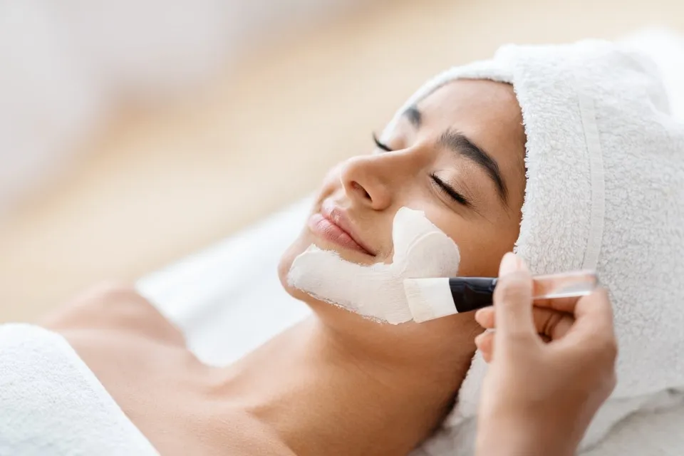 A relaxed woman lying down with a thick white mask applied, receiving facial treatments.