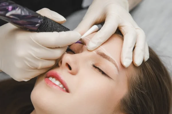 A professional performing a microblading procedure on a female client’s eyebrows using a manual tool to create hair-like strokes in the context of microblading vs micropigmentation